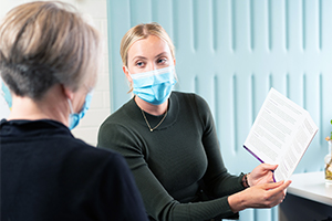 A woman wearing a surgical mask and holding a printed booklet speaks with another woman.