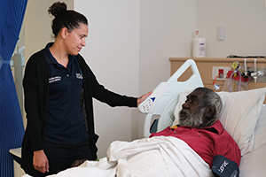 A female Aboriginal Health Liaison Officer stands beside the bedside of an Aboriginal man.