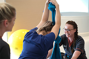 A woman wearing a Fiona Stanley Hospital uniform that reads 'Registered midwife' sits in front of a woman who is squatting on the floor holding onto a birthing silk suspended from the ceiling.