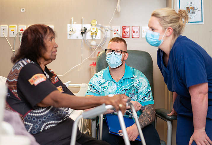 An Aboriginal woman sits on the side of a bed holding the top of a walking frame as she talks with a male and female health professional wearing surgical masks.