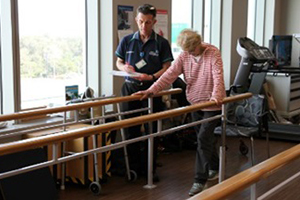 A male physiotherapist stands beside an older woman who is doing physiotherapy exercises.