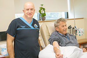 A male enrolled nurse stands beside the bedside of a male patient.