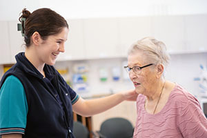 A female physiotherapist stands with her left hand on an older woman's right shoulder