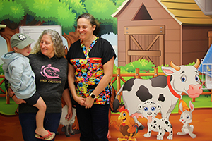 An older woman holds a young boy while a female nurse in colourful scrubs stands beside them in front of a colourful farmyard mural