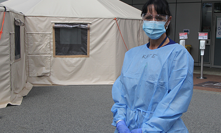 A female nurse wearing a surgical mask, gown, gloves and safety glasses stands outside a tent.