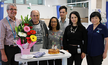 Six people standing in a treatment area. In front of them on a table is some flowers and a cake.
