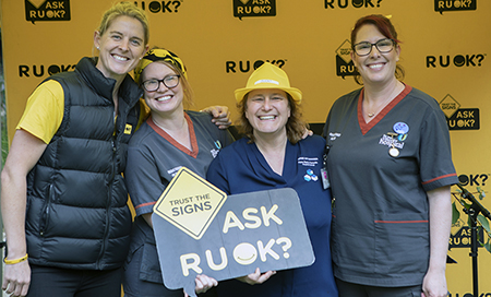 Four woman stand in front of a yellow banner that reads RUOK? One of the women holds a sign that reads Ask RUOK?