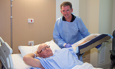 An older man lays in a hospital bed and a male health professional stands beside him.