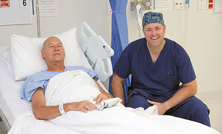 A man sits in a hospital bed. A man wearing theatre scrubs sits next to the bed.