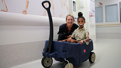A young female nurse sits next to a young female hospital patient who is seated in a colourful toy cart.