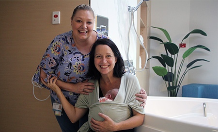 A female midwife stands with a woman who is cradling a newborn baby beside a large bath.