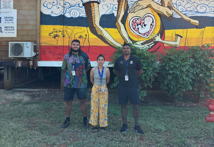 A woman stands flanked by two men in front of a brightly coloured mural